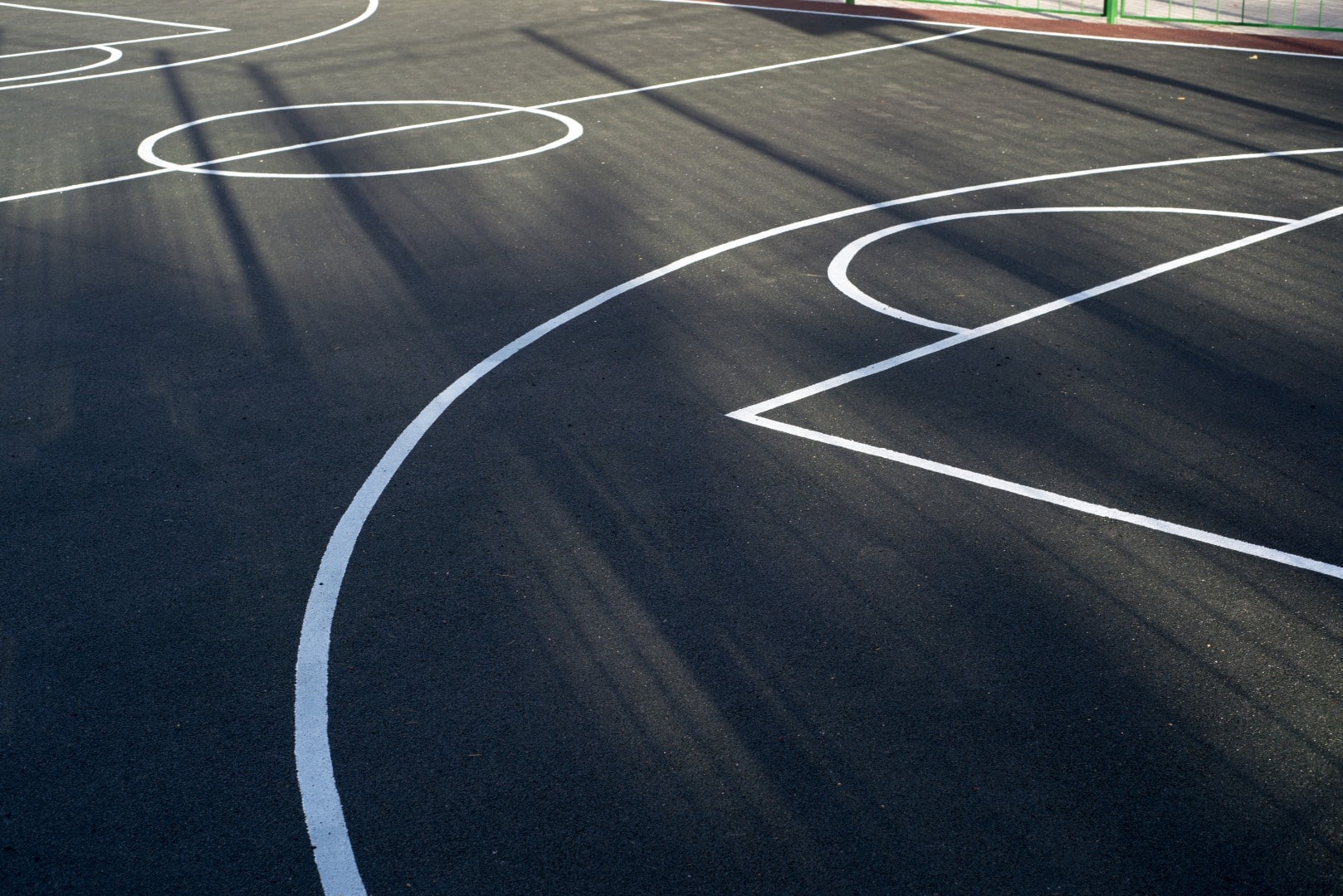 Lignes blanches d'un terrain de basket ou multi-sports noir sous un fort contre-jour projetant des ombres allongées.
